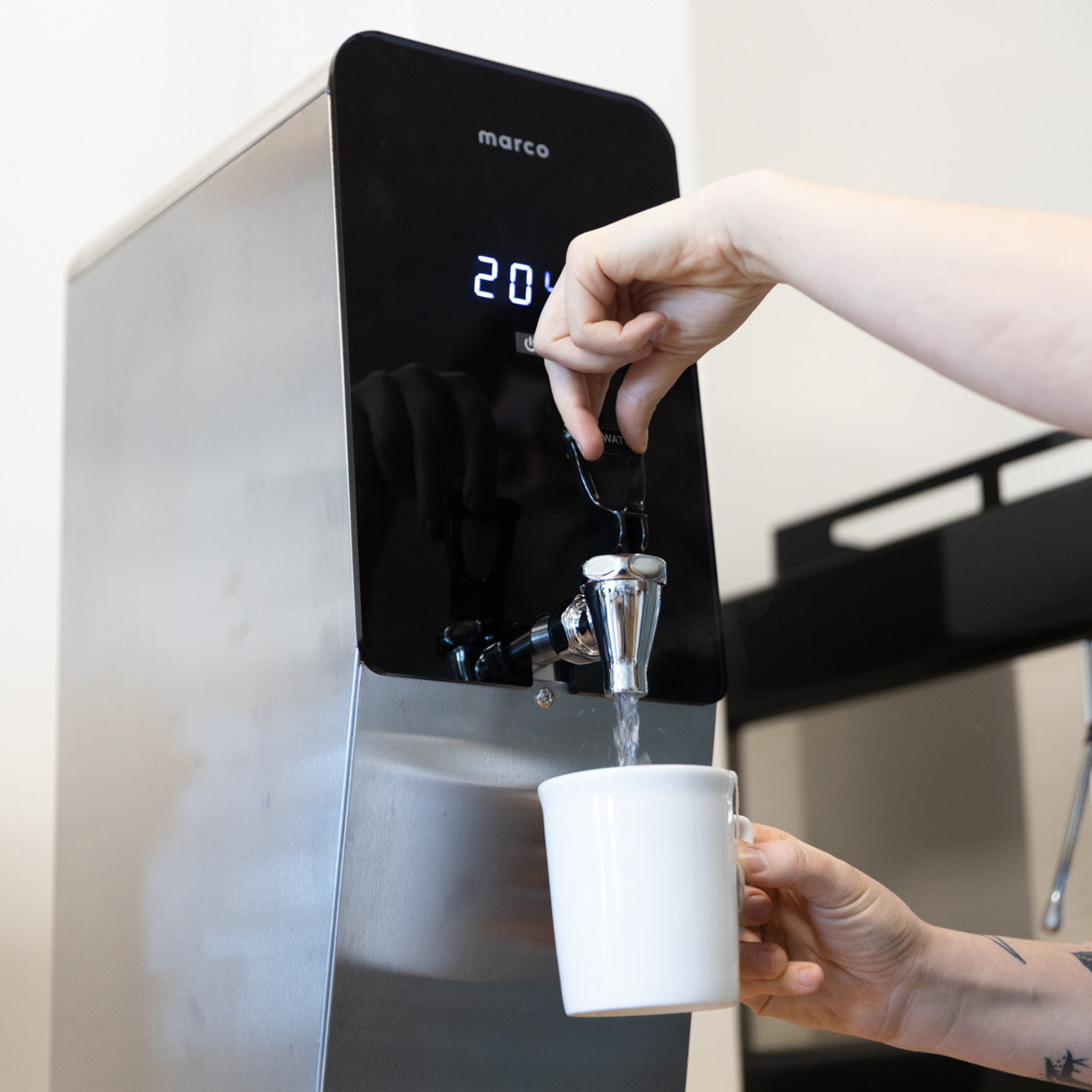 Person filling a white mug with water from a Marco water dispenser.